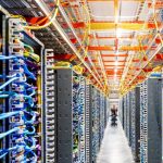 A long, symmetrical aisle inside a high-tech data center featuring rows of server racks with organized, colorful blue and yellow cables, and a technician in the distance