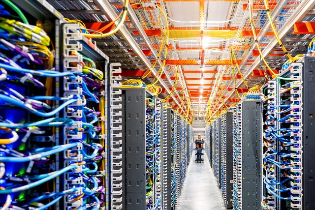 A long, symmetrical aisle inside a high-tech data center featuring rows of server racks with organized, colorful blue and yellow cables, and a technician in the distance