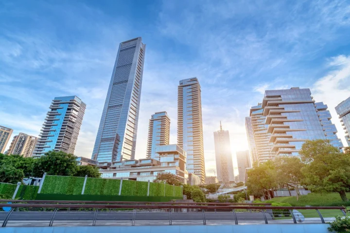 A wide-angle view of modern glass skyscrapers in a sunlit city, featuring a lush green wall and park area in the foreground.